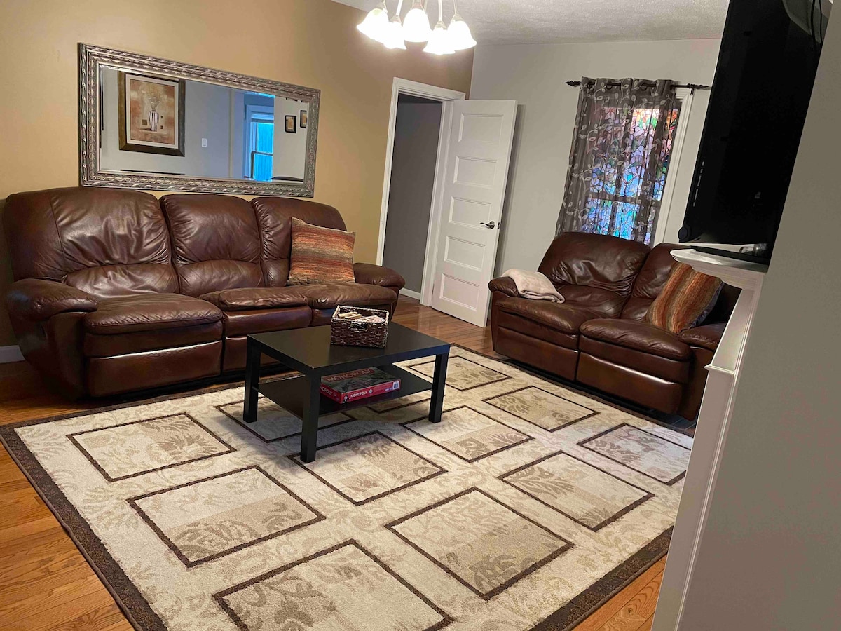 A living room is depicted featuring two chocolate brown leather sofas facing each other. A black coffee table is centered on a cream-colored area rug adorned with geometric patterns. Soft lighting is provided by a chandelier above, complemented by light filtering through sheer curtains.
