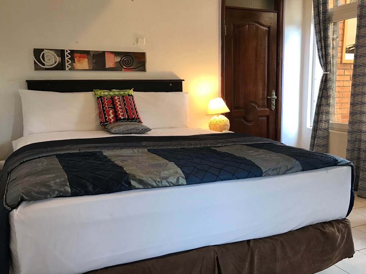 A spacious bed is featured, adorned with a quilted blue coverlet and a decorative pillow. A bedside lamp casts a soft glow, and artwork hangs above the headboard. Natural light filters through a nearby window, complemented by patterned curtains.
