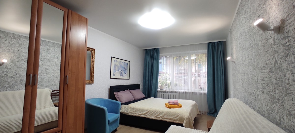 A spacious bedroom features a large bed with a light-colored blanket, accompanied by a comfortable blue armchair. A mirror wardrobe stands against one wall, and a window with blue curtains allows natural light to illuminate the room.