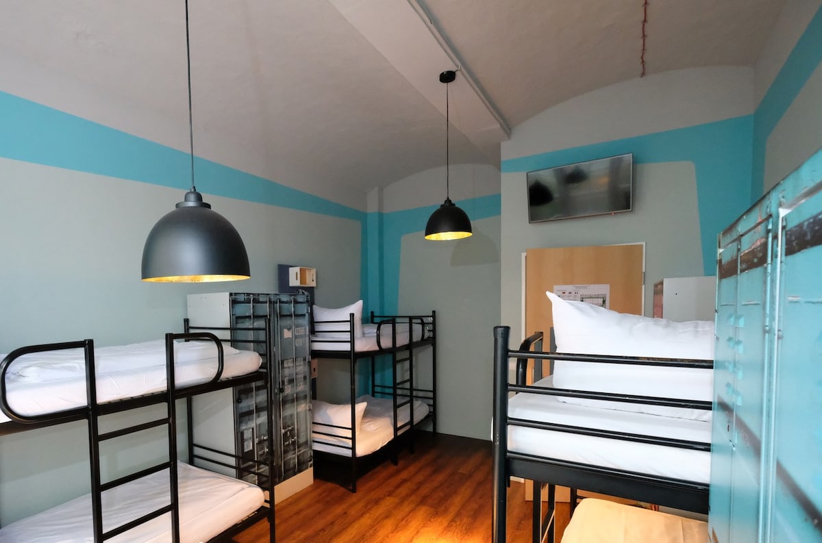 A shared dormitory space features multiple bunk beds arranged against the walls, providing various sleeping options. The room is illuminated by pendant lights hanging from the ceiling. Light wood flooring complements the vibrant blue accents on the walls, while a television is mounted at the far end.