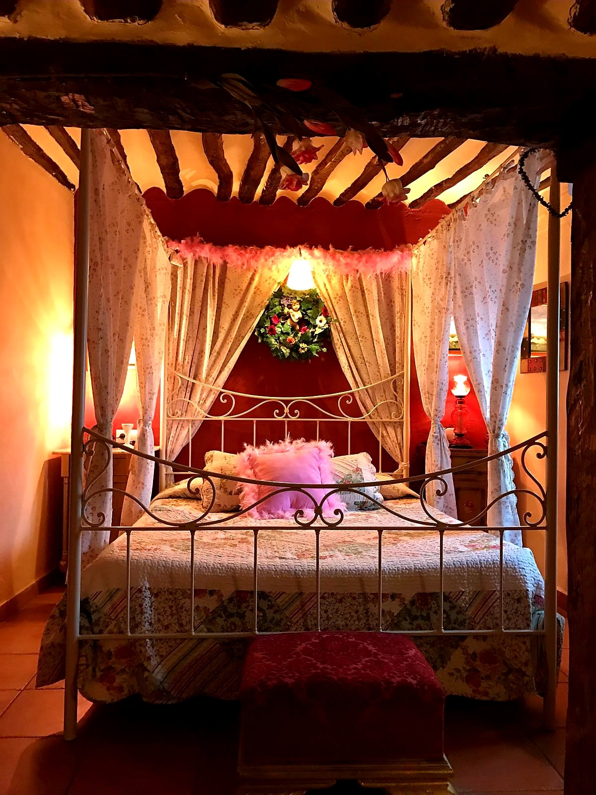 A cozy bedroom features a four-poster bed draped in soft, flowing fabrics. Warm lighting creates a gentle ambiance, while floral patterns adorn the bedding. Rustic wooden beams enhance the room's charm, complemented by decorative elements such as a hanging wreath and bedside lamps.