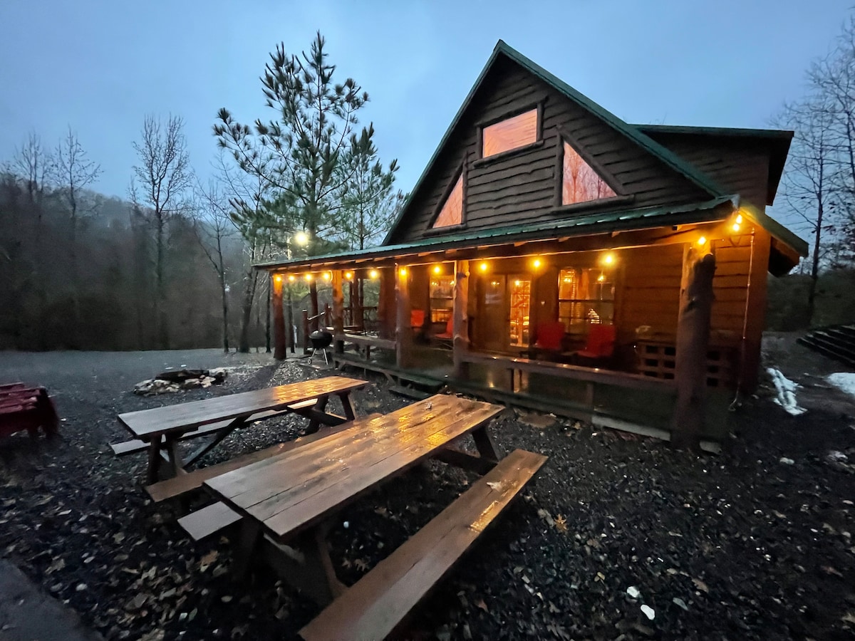 The cabin exterior features warm string lights illuminating a large covered porch. Two picnic tables are arranged on the gravel ground, with a fire pit nearby. Trees surround the area, adding to the rustic charm in the twilight ambiance.