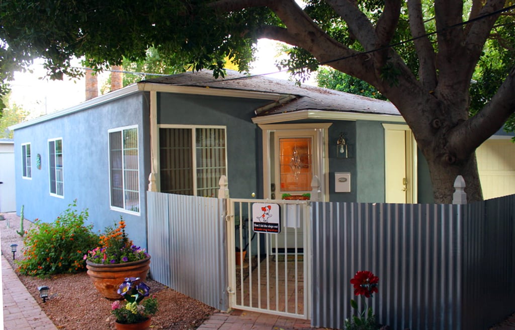 The exterior of the casita is featured, showcasing a blue painted façade with large windows. A decorative fence surrounds a shaded entrance, complemented by vibrant flower pots and foliage. A welcoming sign indicates dog-friendly amenities, enhancing the inviting outdoor space.