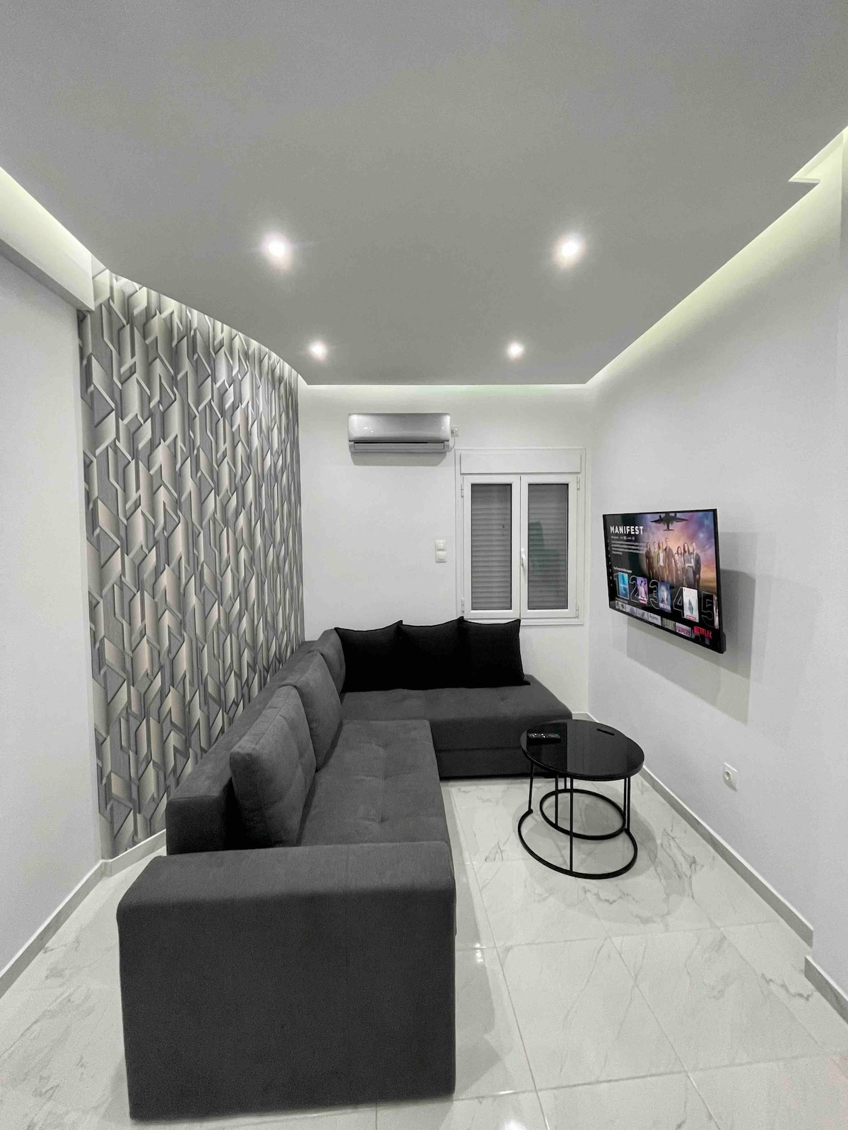 A modern living area features a grey sectional sofa complemented by black cushions. A small round table is positioned in front, and a flat-screen television is mounted on the wall. Natural light enters through a window with white blinds, while recessed lighting highlights the neutral design.