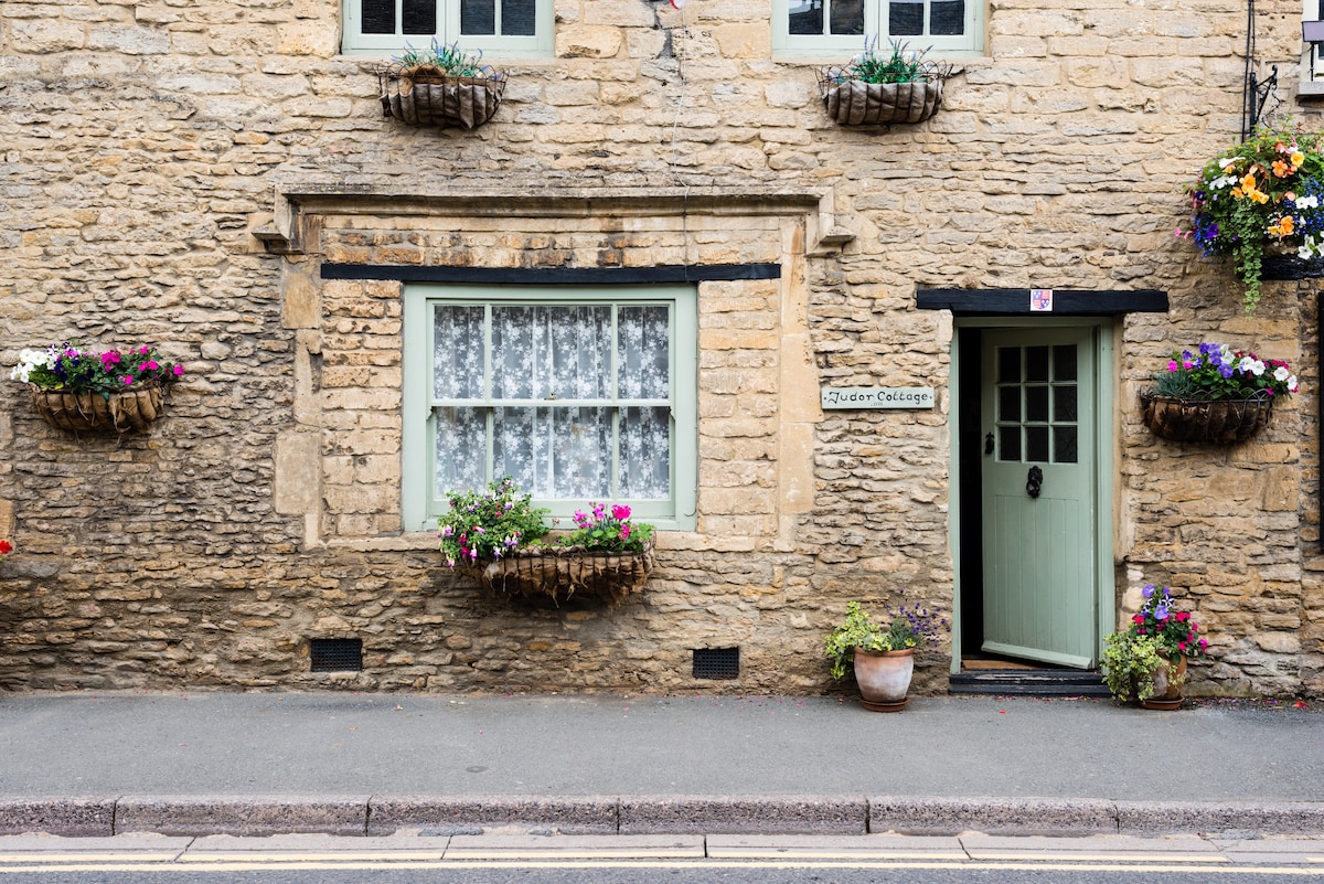 The charming exterior of Tudor Cottage features Cotswold stone walls adorned with window boxes filled with colorful flowers. A decorative front door, framed by a simple entrance, complements the inviting facade. A lace-curtain-adorned window enhances the cottage's traditional character.