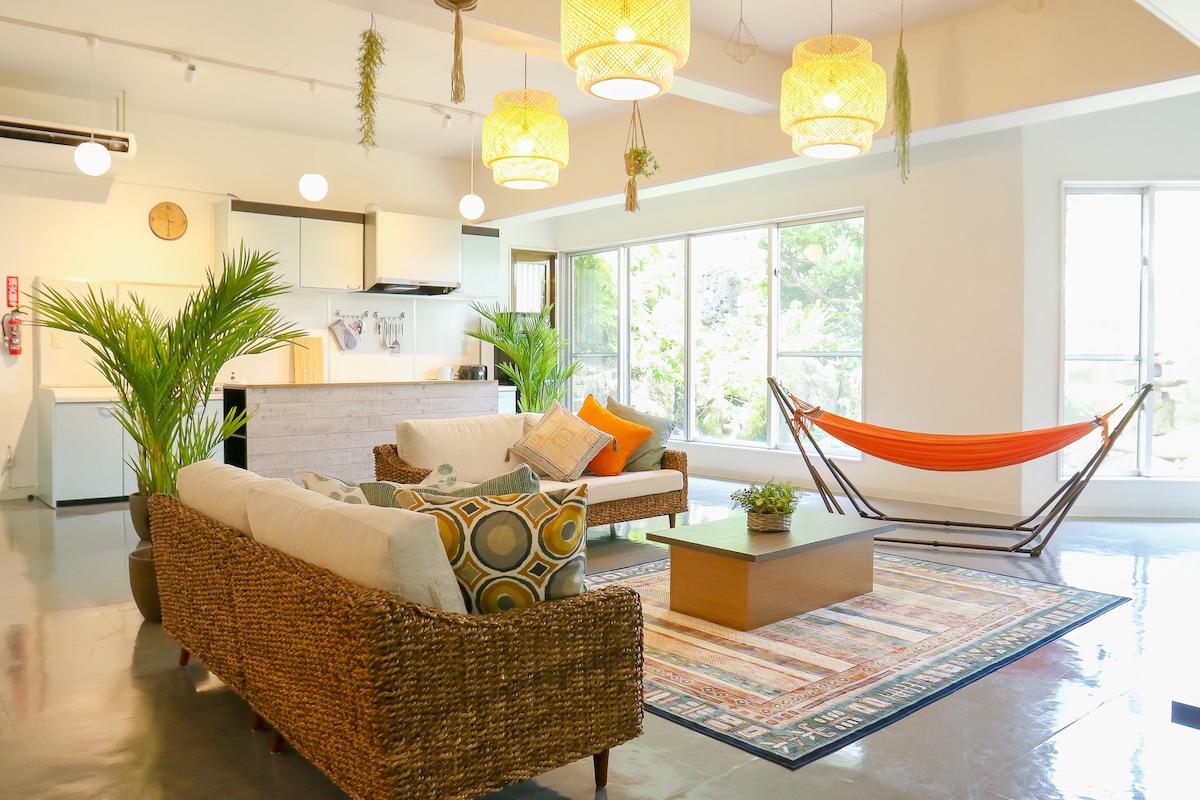 The open-plan living area features comfortable seating with neutral-toned couches accented by colorful cushions. A central coffee table sits atop a patterned rug. Large windows allow natural light to fill the space, complemented by hanging light fixtures and indoor plants, creating a relaxed environment.