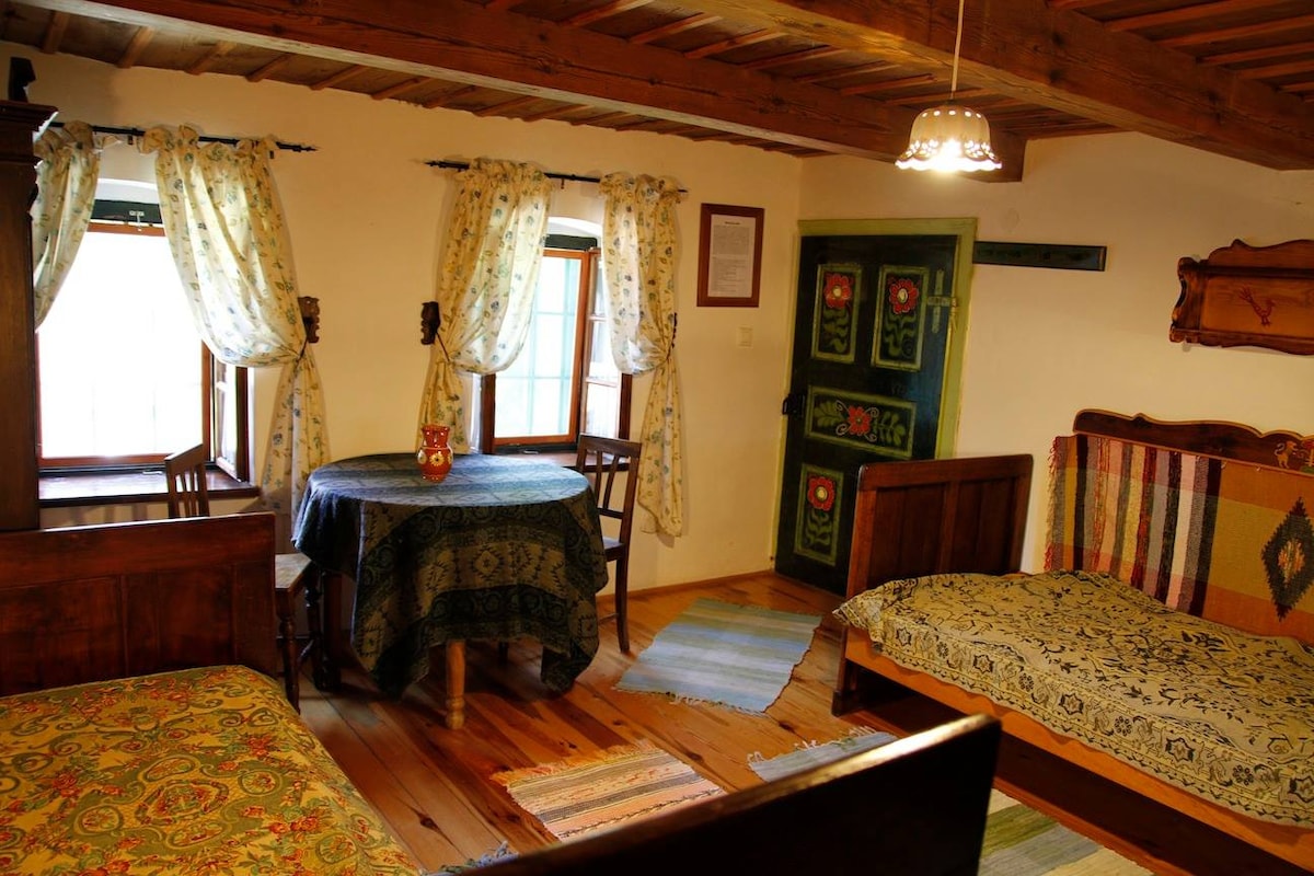 The room features traditional wooden furnishings, including two beds adorned with patterned beddings. A round table with chairs sits centrally, surrounded by bright floral drapes framing two windows. A rustic-style cabinet adds character to the space, complemented by wooden beams overhead.