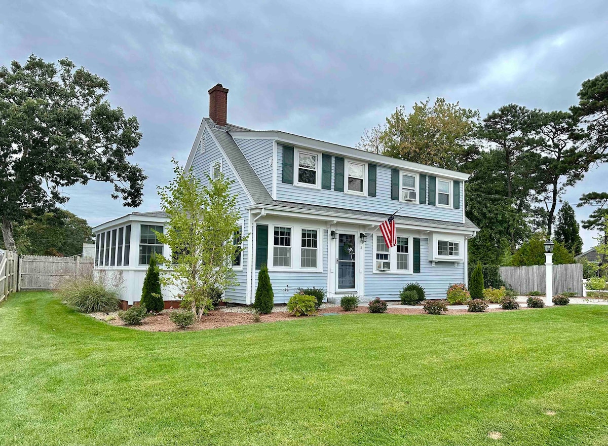 A charming two-story home features light blue siding and green shutters, complemented by neatly manicured landscaping. An American flag hangs by the front door, while a spacious yard provides an inviting outdoor setting. The sun room on the side enhances the overall appeal.