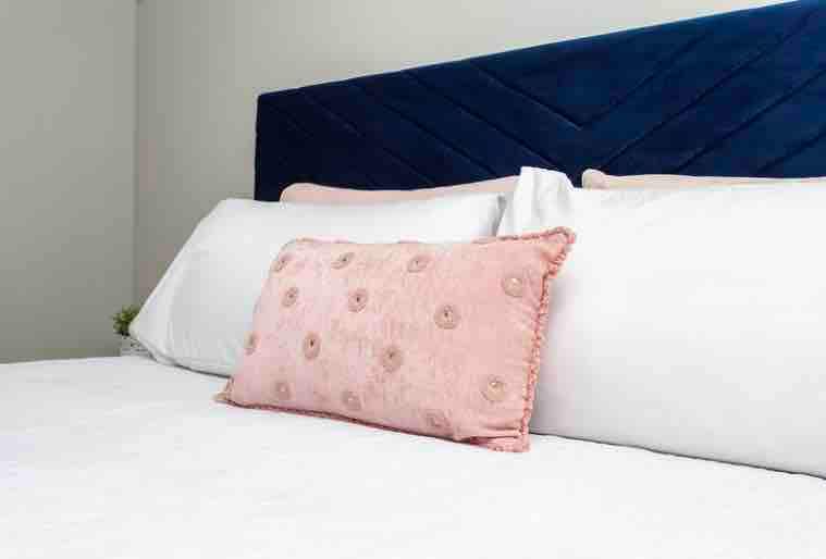A spacious bed is positioned against a textured navy blue headboard, creating a calming focal point. Soft white bedding is arranged neatly, while a decorative pink pillow with circular embellishments adds a touch of color and comfort, inviting relaxation.