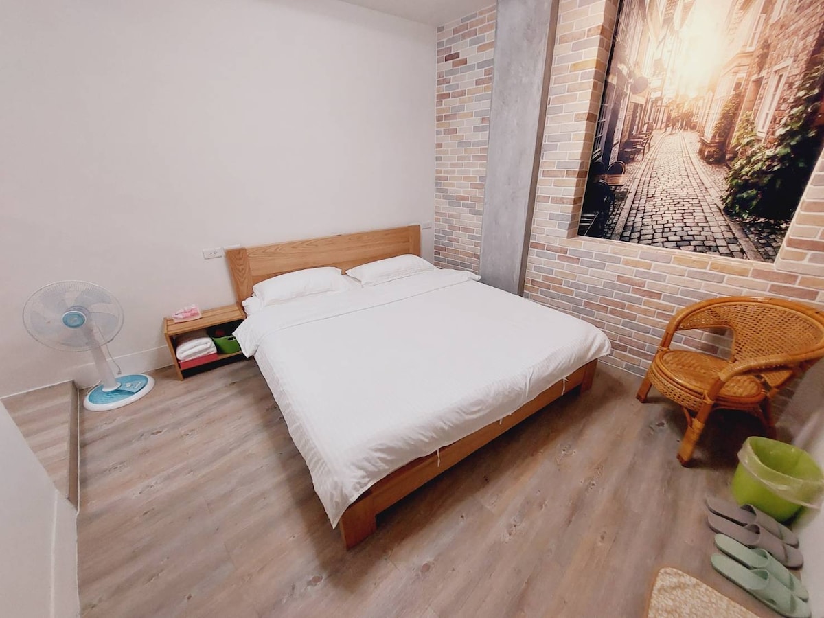 A minimalist bedroom features a spacious bed against a textured wall. A decorative photo of a cobbled street adds interest. A rattan chair and a fan are positioned nearby, with soft lighting highlighting the room's clean lines and wooden accents.