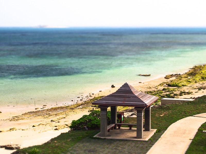 A shaded gazebo is positioned along a serene stretch of beach, with gentle waves lapping at the shore. The coastline showcases a blend of sand and rocky formations, while clear blue waters extend toward the horizon, inviting relaxation and tranquility.