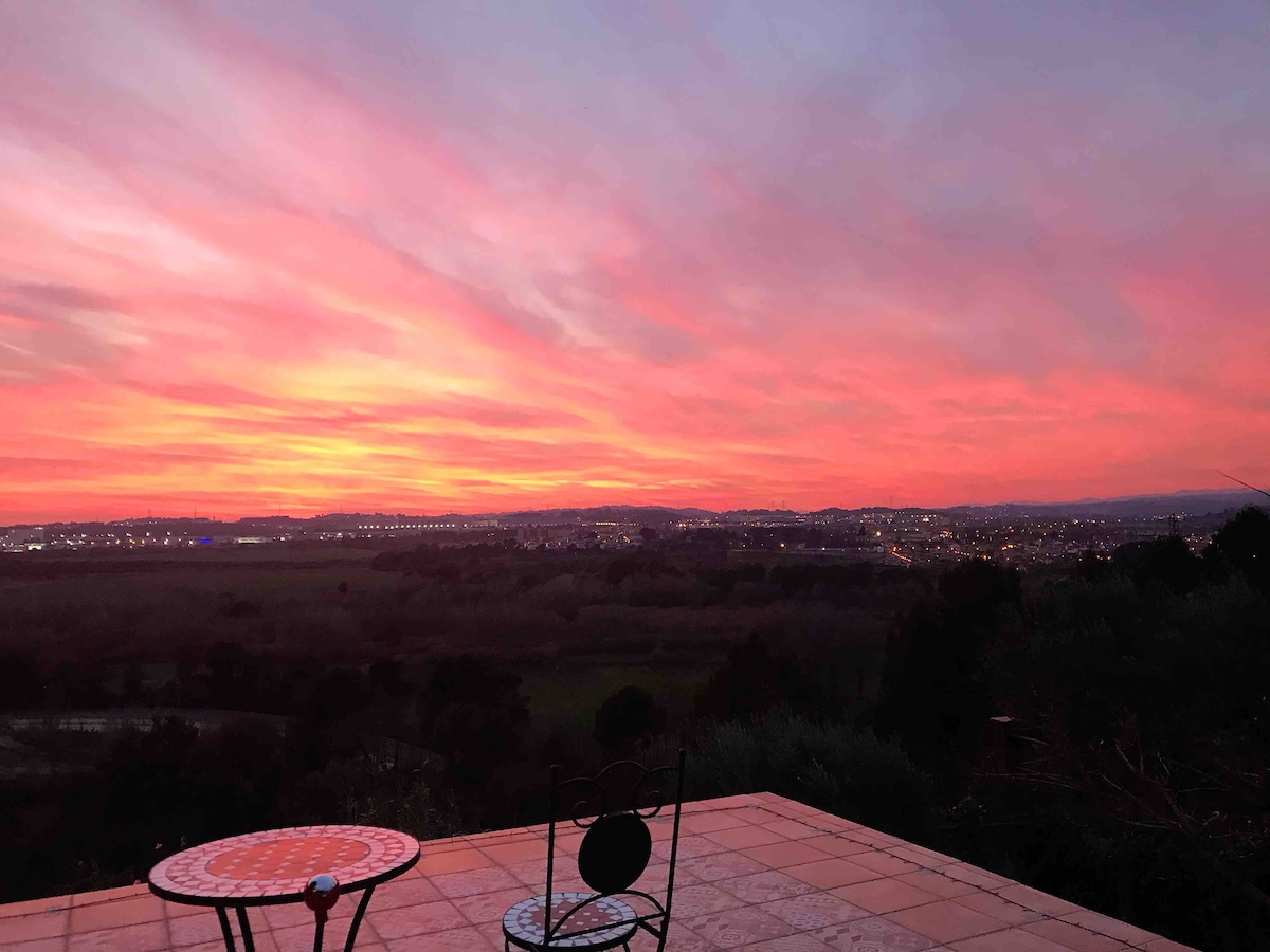 A serene outdoor terrace is featured, showcasing a round table and a black chair. A vibrant sunset paints the sky in shades of pink and orange, casting a warm glow over the scenic landscape and distant city lights below.