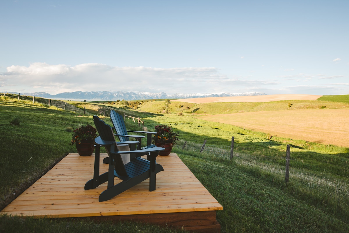A wooden deck features two comfortable Adirondack chairs overlooking expansive rolling hills and fields. Potted flowers add a touch of color, while the distant mountain range is visible against a clear sky, creating a peaceful outdoor setting.