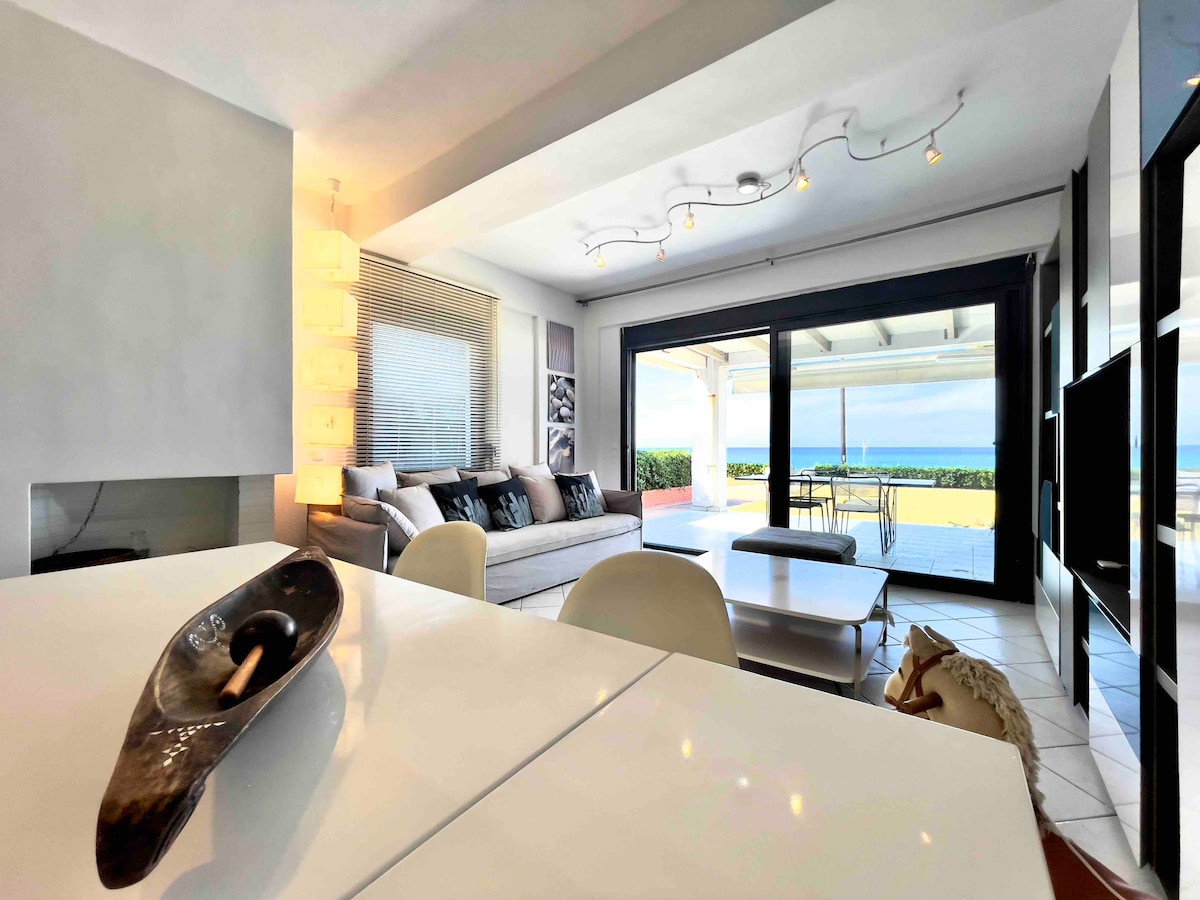 A modern living space is seen with contemporary furniture, including a spacious sofa and a stylish coffee table. Large glass doors provide a view of the outdoor area and the coast, allowing natural light to fill the room. Subtle decorative accents enhance the design.