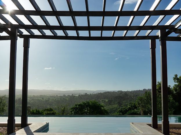 A wooden pergola frames a serene view of rolling hills under a clear blue sky. The sun casts a soft light on the area surrounding a tranquil swimming pool, enhancing the sense of relaxation and openness.