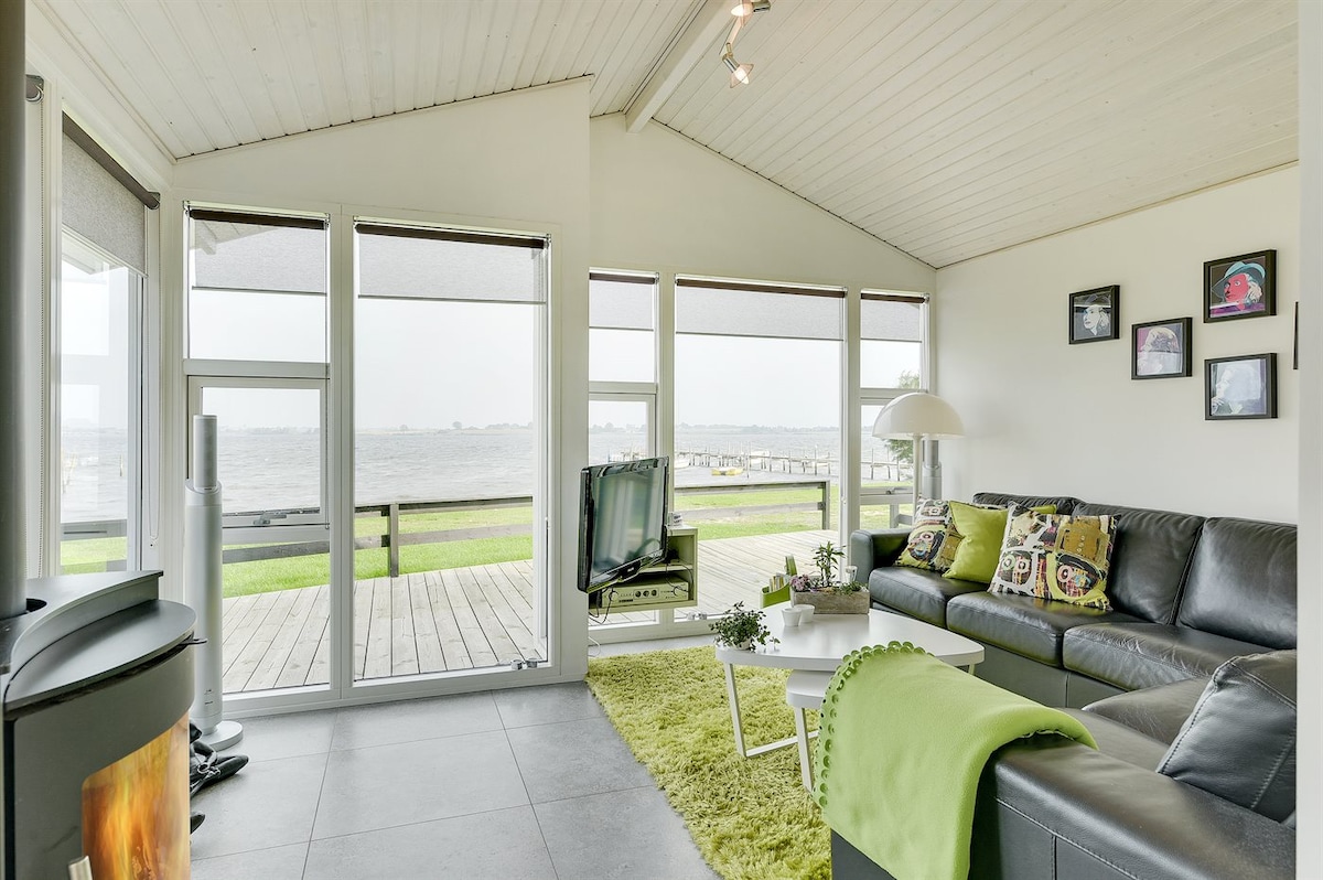 A spacious living area features large windows providing views of the water. A sleek black sectional sofa is accompanied by decorative pillows, while a green area rug adds a touch of color. A modern television is positioned for easy viewing, with a cozy fireplace nearby.