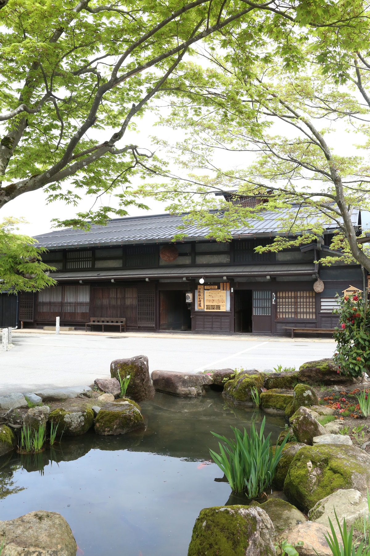 A traditional wooden building with a sloped roof is showcased amidst a landscaped garden. A tranquil pond reflects the structure, surrounded by stones and greenery. The entrance is highlighted by a wooden sign, inviting guests to explore the serene surroundings.