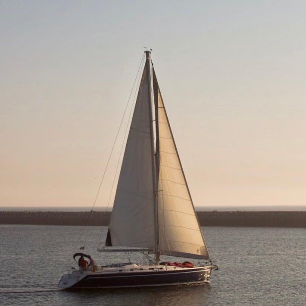 A sailboat glides gracefully across calm waters, with its large white sail billowing in the gentle breeze. The serene atmosphere is enhanced by the soft light of the setting sun, casting a warm hue over the scene.