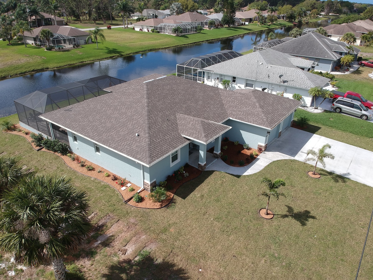 The property features a single-story home set in a lush green landscape, surrounded by a manicured lawn and palm trees. It is positioned near a tranquil waterway, with neighboring residences visible in the background.