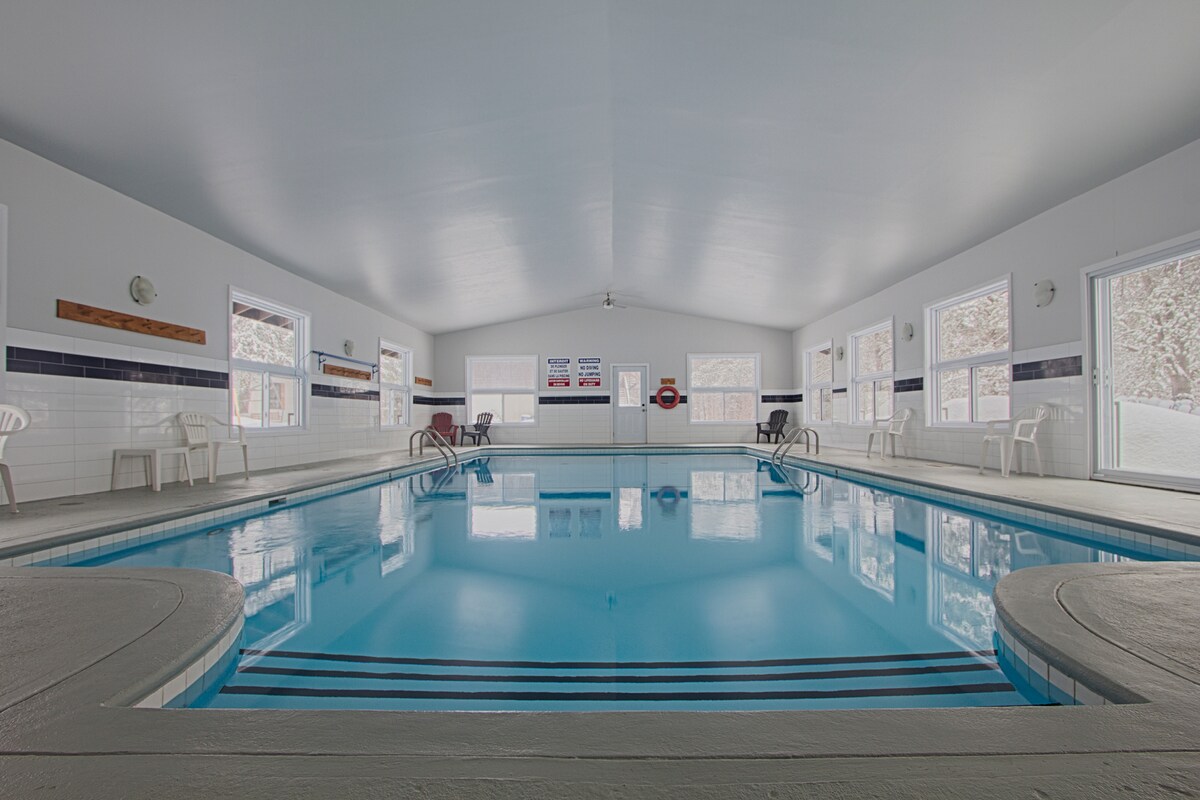 An indoor pool area is showcased with clear blue water reflecting natural light from multiple large windows. White walls and ceiling enhance the bright atmosphere. Several white plastic chairs are arranged beside the pool, providing seating for relaxation.