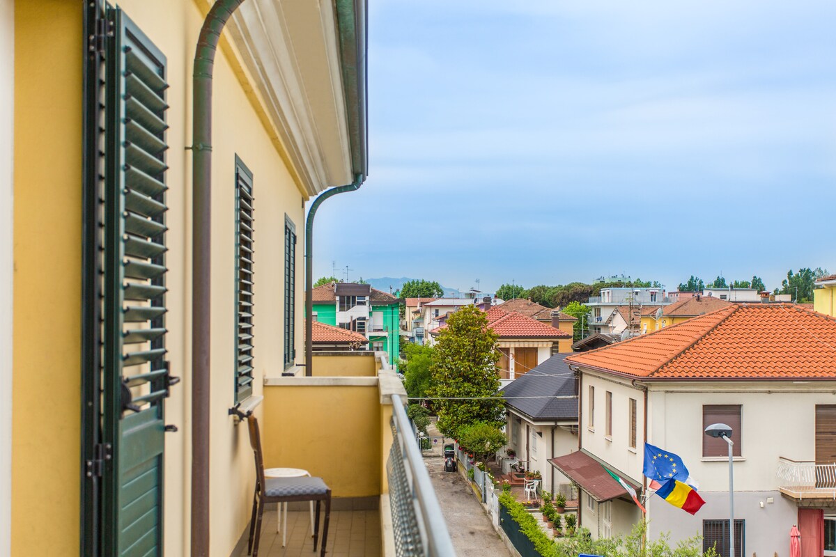 A balcony view features colorful residential buildings and rooftops in the distance. A small chair is positioned on the balcony, providing a spot to enjoy the scenery. Lush greenery and trees can be seen along the street, creating a vibrant neighborhood atmosphere.