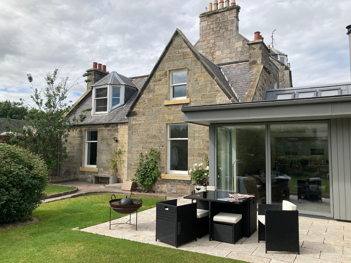 The exterior of the cottage features a blend of traditional stonework and modern extension. The walled garden is landscaped with a manicured lawn. A patio dining area with stylish furnishings is visible alongside a BBQ fire pit, creating a perfect space for outdoor gatherings.