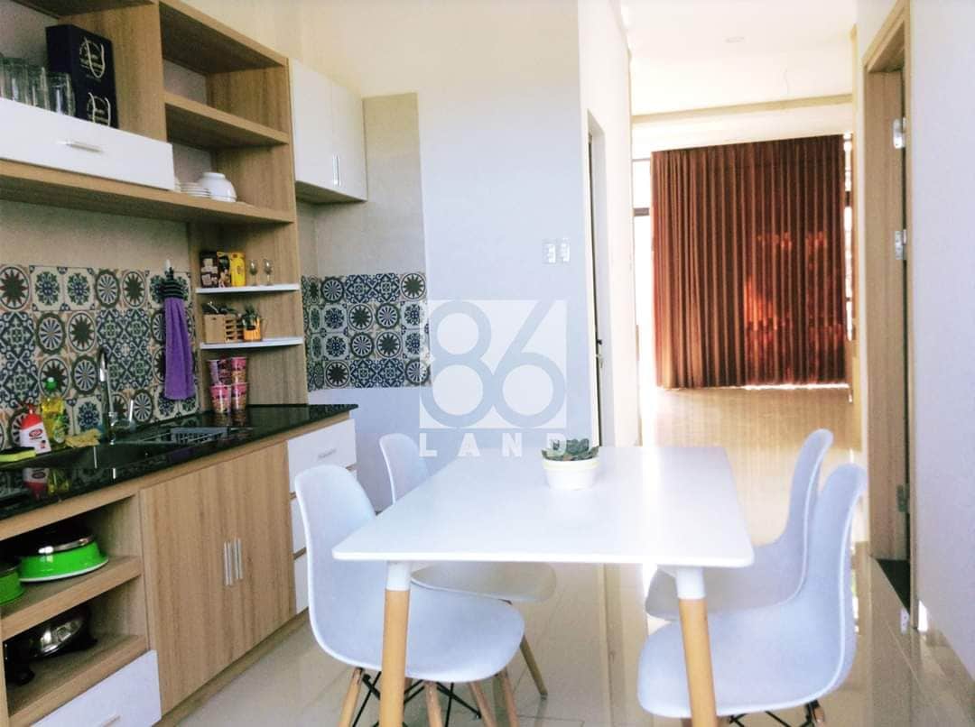 A modern kitchen area features light wooden cabinetry and patterned tiles. A white table is accompanied by four minimalist chairs. Natural light is allowed in through large windows, complementing the airy atmosphere.