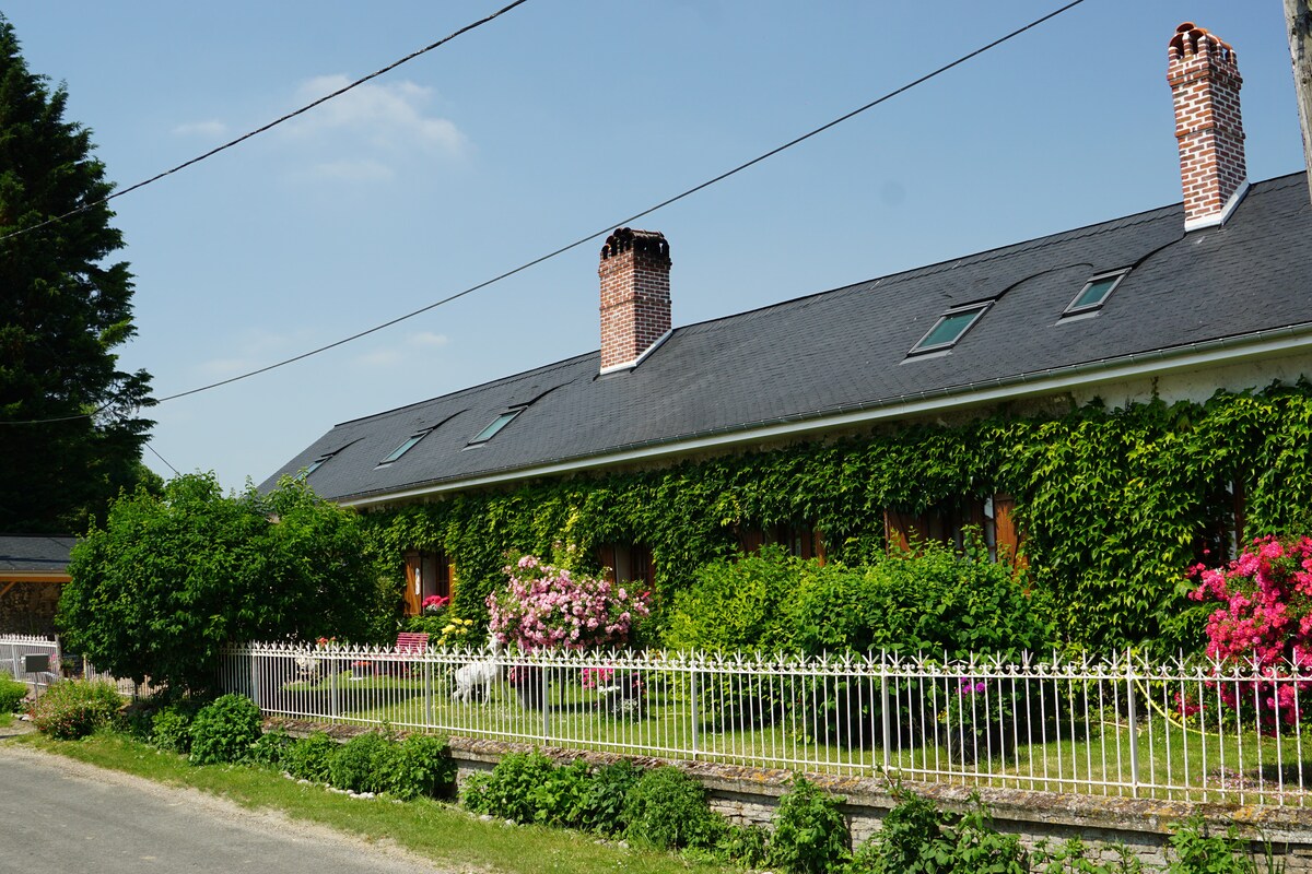 A charming building is framed by lush greenery, with vibrant flowers adding color to the façade. Two prominent chimneys rise from the roof, enhancing the countryside appeal. A white picket fence runs along the front, contributing to the serene and welcoming atmosphere.