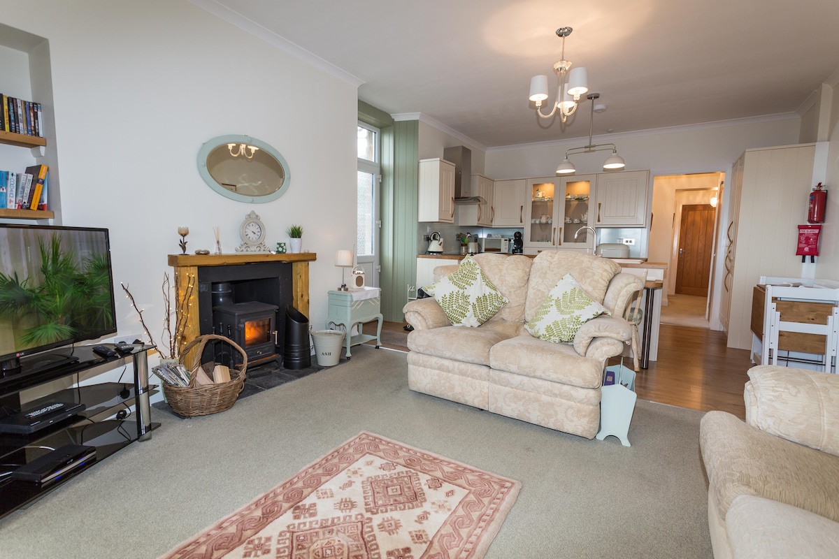 An open-plan living area features a light-colored, patterned sofa and armchair, positioned near a wood burner and a coffee table. A television is situated on a stand, while shelves filled with books add to the decor. Warm light illuminates the space, complementing the neutral décor.