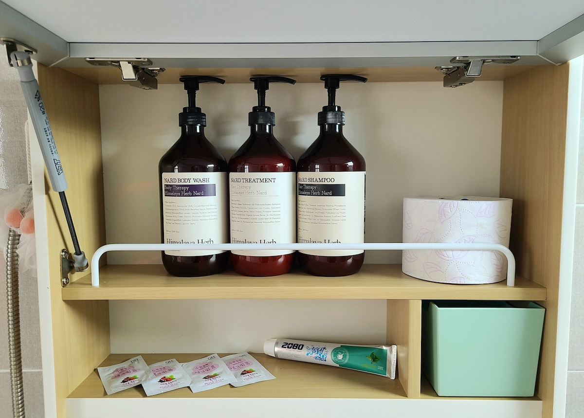 A well-organized bathroom shelf displays three large bottles of body wash, shampoo, and treatment, all labeled clearly. A roll of toilet paper and a green storage box are also present, emphasizing the functional and accommodating design of the space.