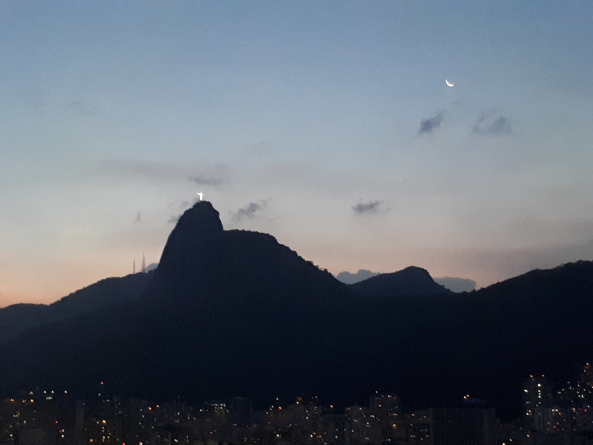 The image captures a scenic view of the iconic Cristo Redentor statue atop a mountain at dusk, with a crescent moon visible in the sky. The surrounding landscape features silhouettes of surrounding mountains and a panoramic cityscape illuminated by soft evening lights.