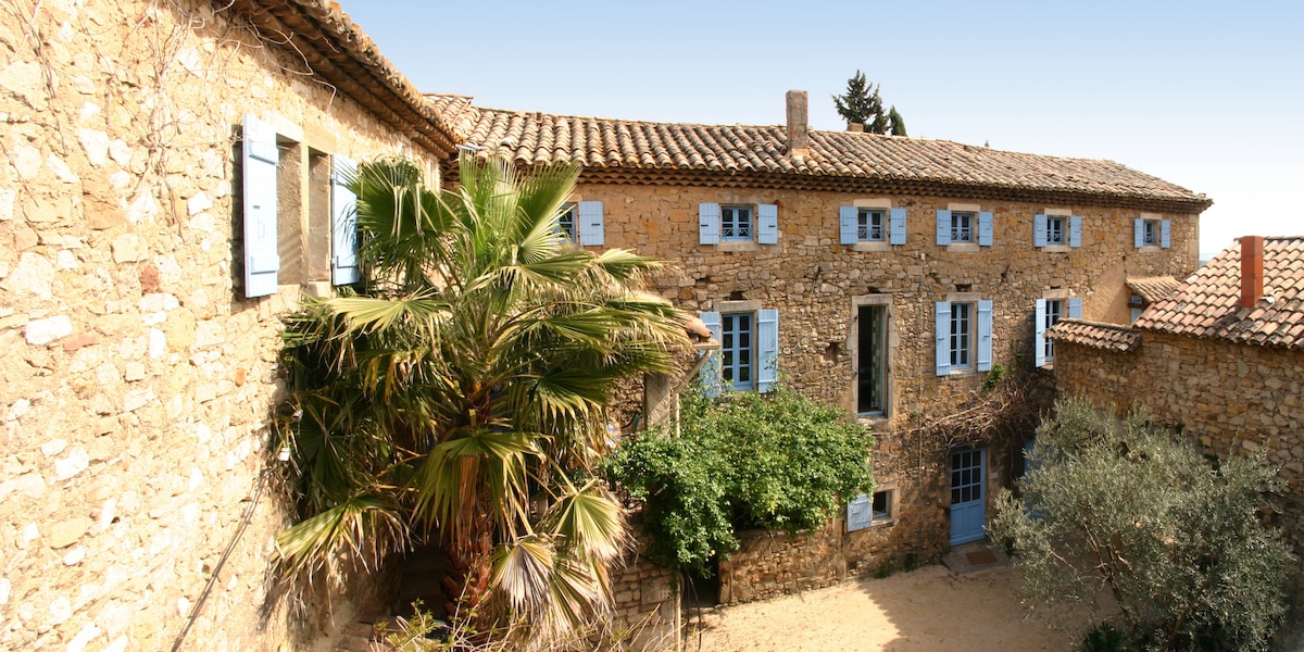A historic stone building of the 17th century is showcased, featuring blue window shutters that contrast with the natural stone facade. Lush greenery, including a palm and olive trees, surrounds the courtyard, creating a serene outdoor space illuminated by soft daylight.