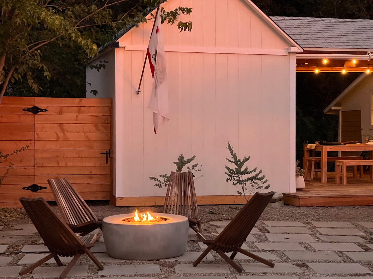 An outdoor fire pit is surrounded by four wooden lounge chairs on a stone patio, offering a cozy gathering space. A small white structure is visible in the background, with warm lighting illuminating the area. Nearby, a dining space can be seen on the deck.