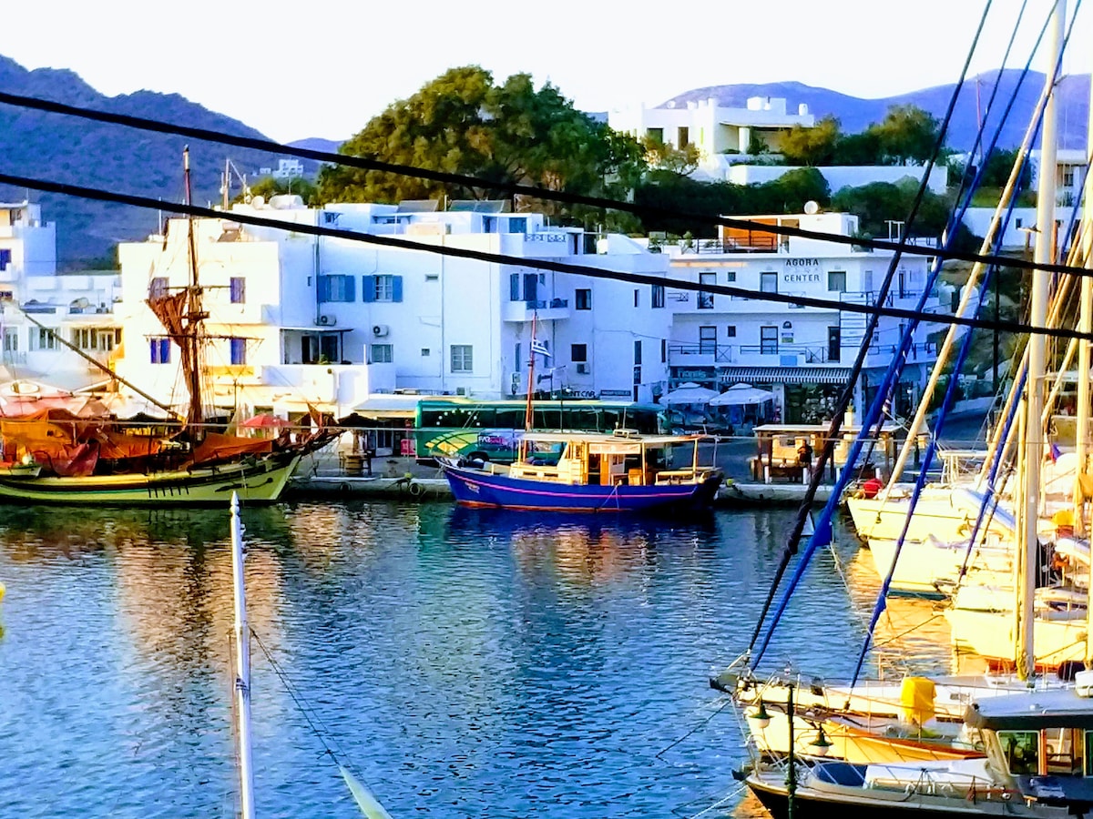 Vibrant fishing boats and yachts are anchored in the calm waters of the port. The shoreline features white buildings with blue accents, reflecting the serene ambiance of the area. Mountains rise in the background, providing a scenic contrast to the waterfront.