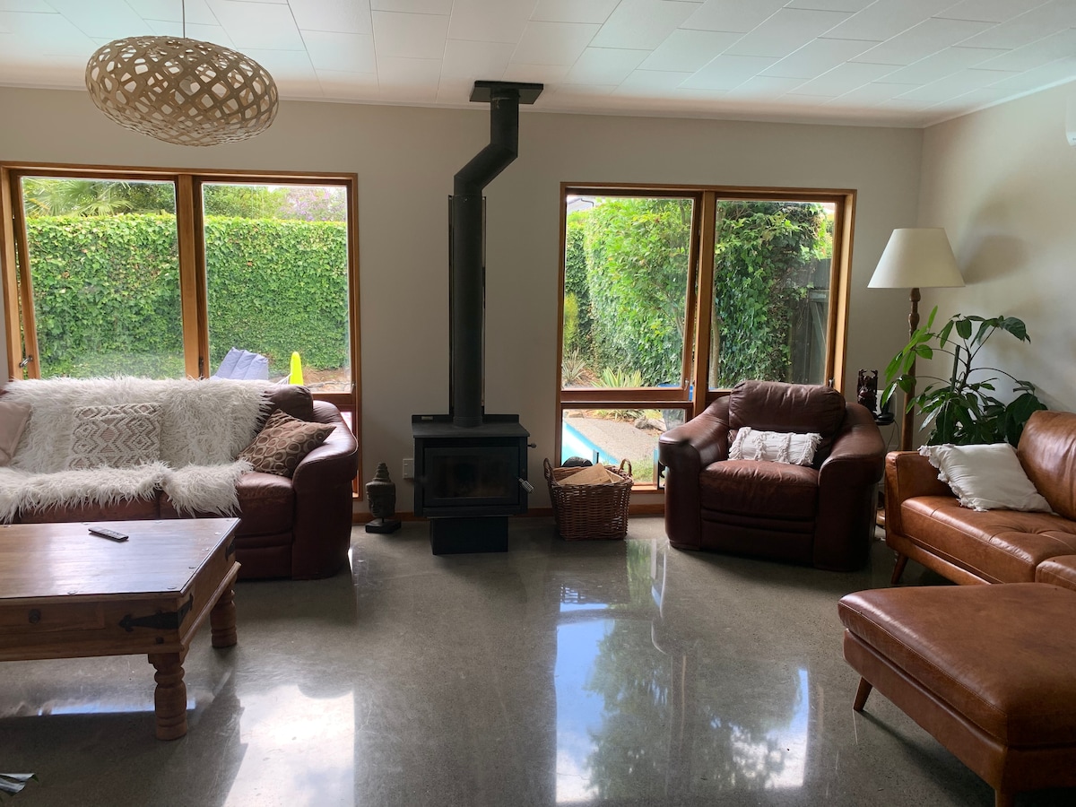 A bright living area features two leather sofas positioned around a wooden coffee table. Large windows provide views of the garden, with greenery visible outside. A modern wood stove is centrally located, and a floor lamp adds light near a floor plant in the corner.