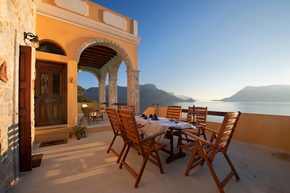 An outdoor dining area features a wooden table surrounded by several chairs, all positioned on a terrace. The scene overlooks a serene seascape with distant hills, bathed in warm sunlight. Architectural details include arched doorways and stone accents on the structure.
