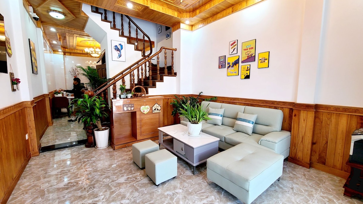A bright living space features a light blue sectional sofa with decorative cushions, complemented by a grey coffee table. Potted plants enhance the area, while framed artwork adds a splash of colour to the walls. A wooden staircase leads to the upper floor.