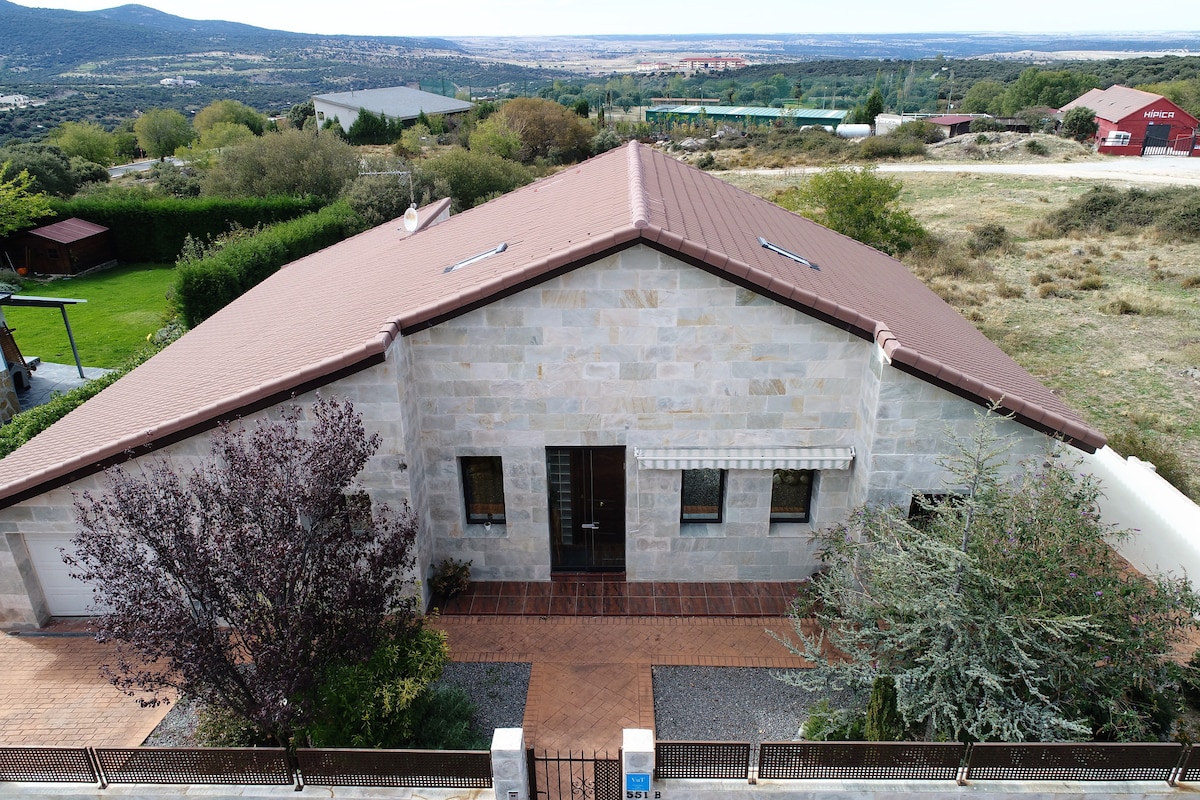 The image presents an aerial view of a spacious stone house with a brown roof. Surrounding greenery and a clear sky are visible, along with distant hills. The entrance features a double door, flanked by windows that allow natural light to enter.