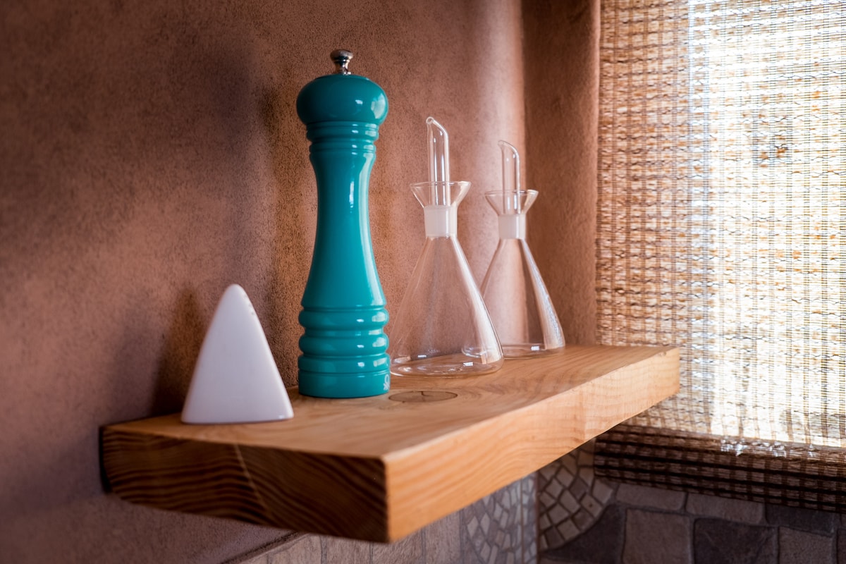 A wooden shelf displays a turquoise pepper grinder alongside glass oil and vinegar dispensers. A white ceramic triangle is positioned to the left. Natural light filters through a woven window covering, illuminating the warm tones of the wall.