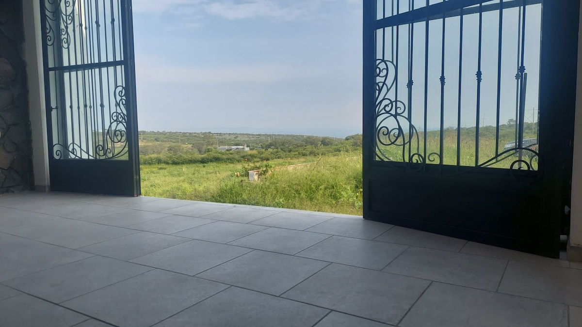 Two large wrought iron doors are open, providing a view of a lush green landscape stretching towards the horizon. The floor is tiled in neutral colors, reflecting natural light and enhancing the spacious feel of the entryway.