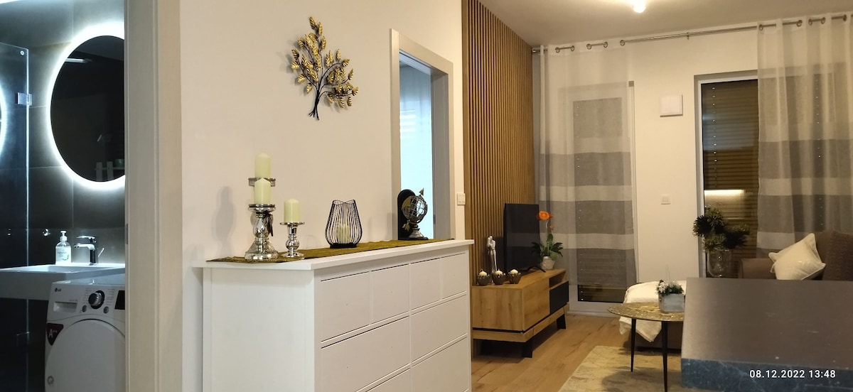 A modern living space features a sideboard adorned with decorative items and candles. A cozy seating area displays a neutral-toned couch and a wooden coffee table. In the background, a well-lit bathroom is visible, highlighted by a circular mirror, while large windows allow natural light to fill the room.