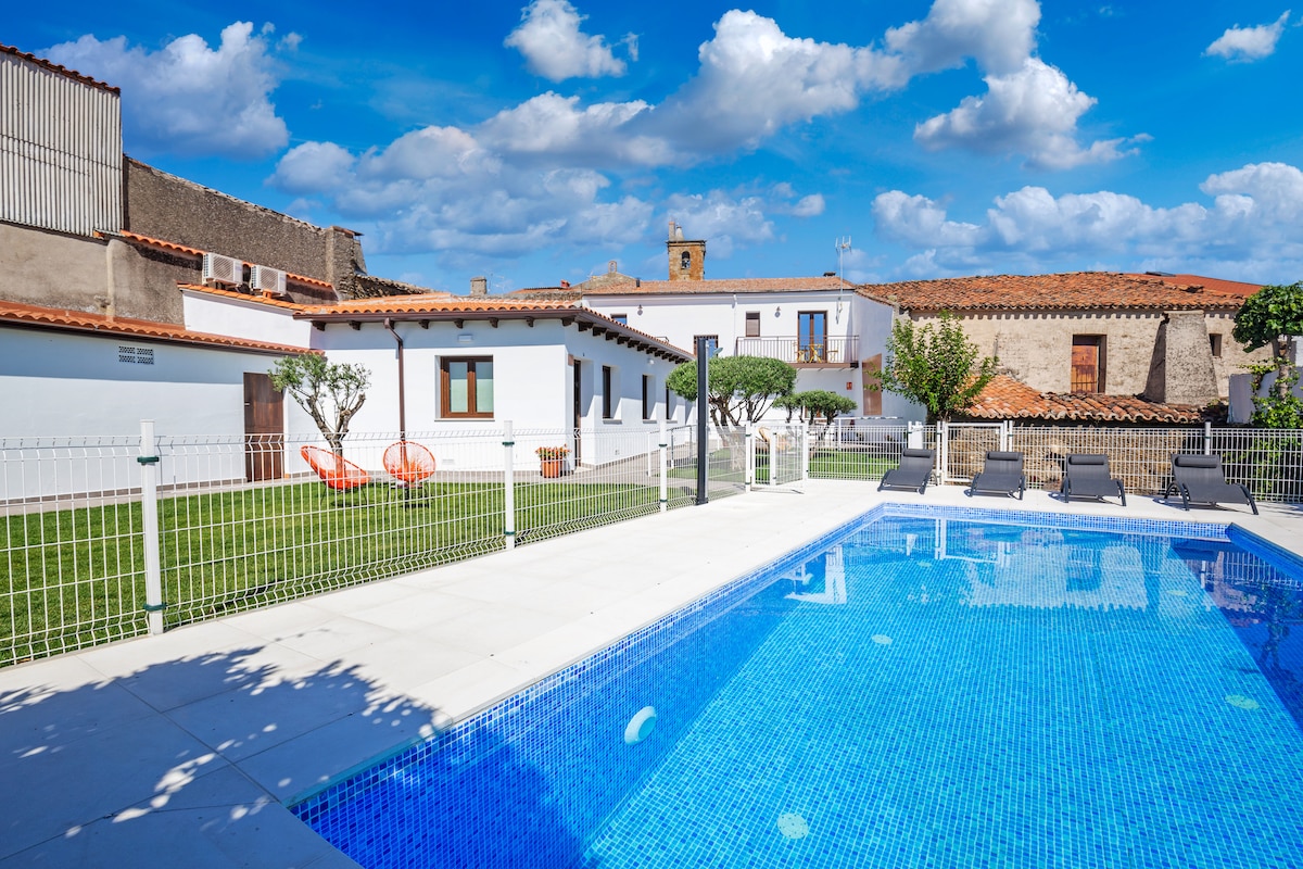 A well-maintained outdoor area features a sparkling swimming pool surrounded by loungers. Lush green lawns host neatly trimmed shrubs and trees. The building presents a modern design, with windows allowing natural light. A clear sky with scattered clouds enhances the serene atmosphere.