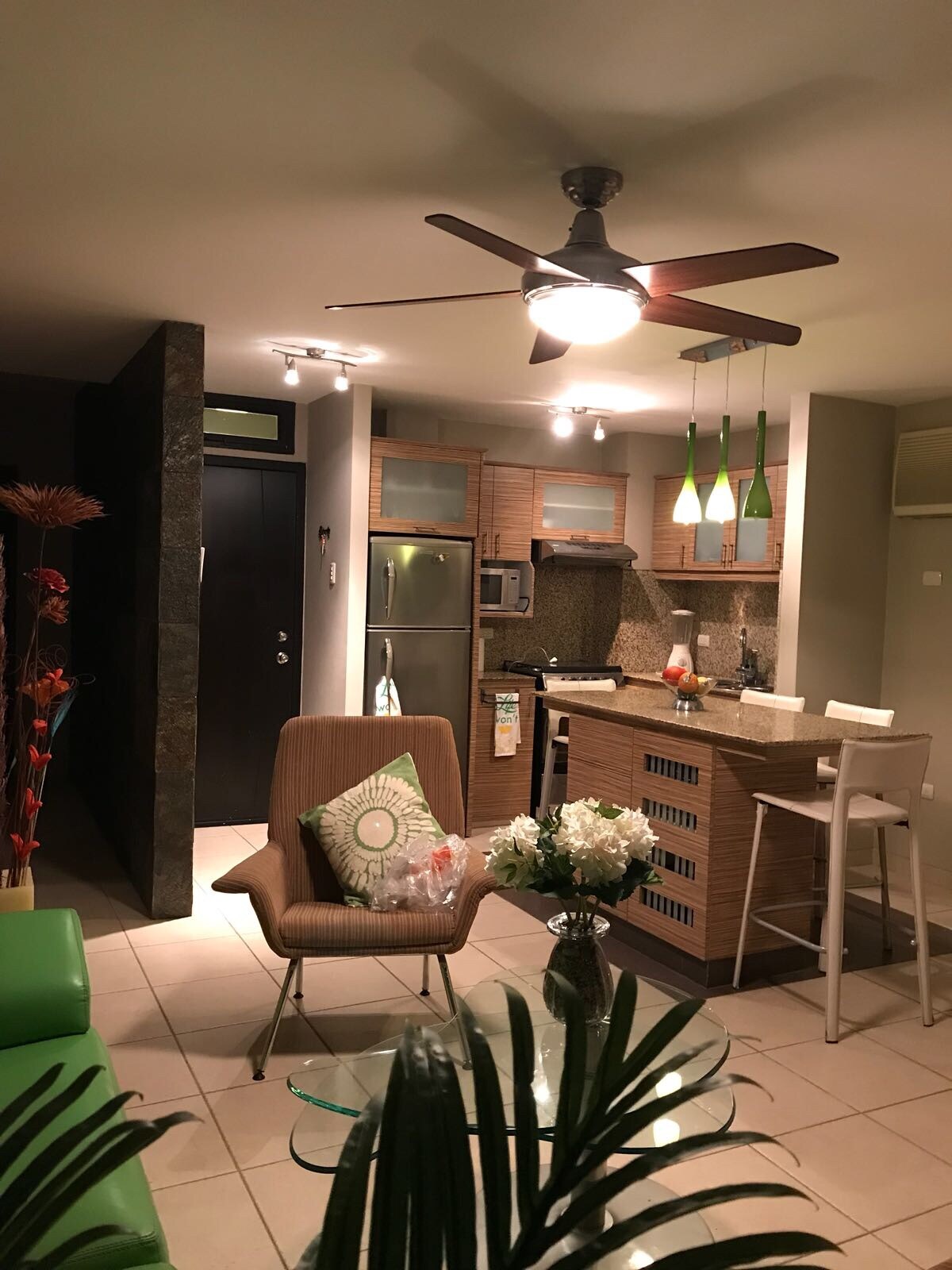 A modern kitchen featuring wooden cabinetry and stainless steel appliances is illuminated by pendant lights. A comfortable seating area with a green chair and a glass table is visible, accompanied by a vase of white flowers. The room's tile flooring complements the contemporary design.