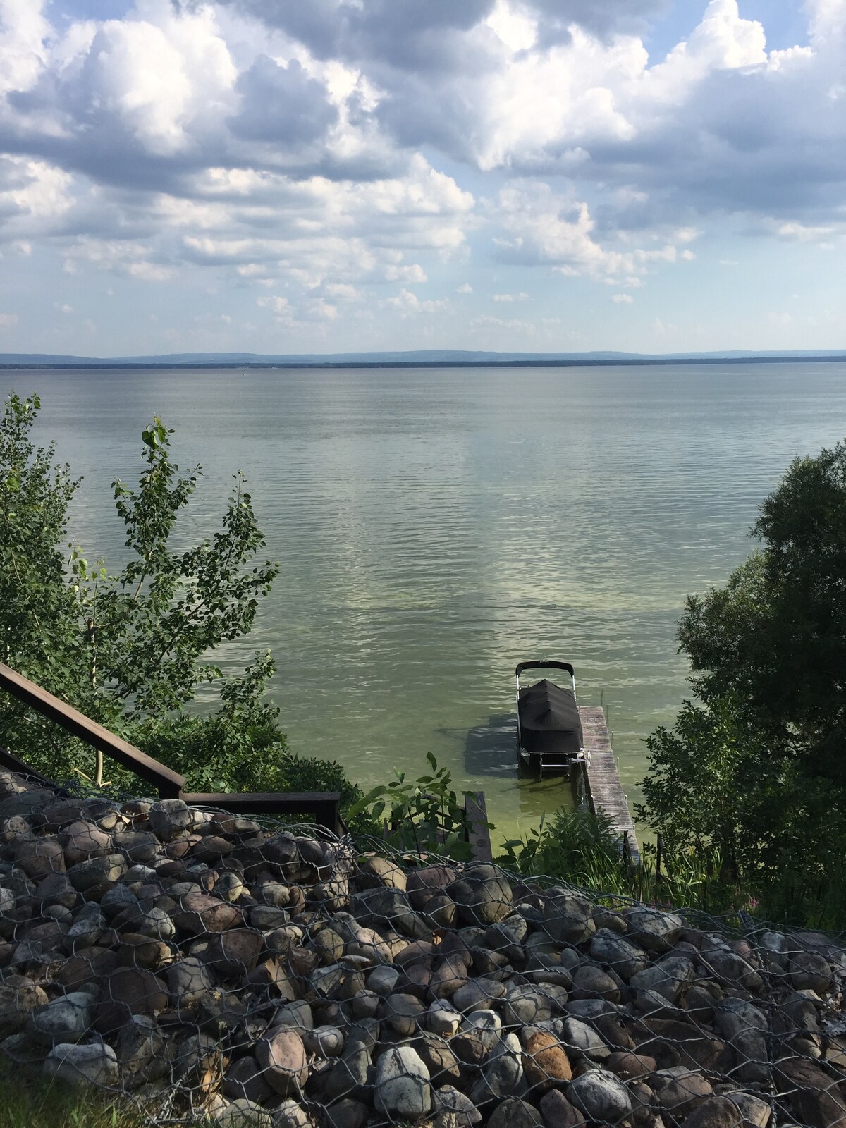 A view captures the serene Oneida Lake, with calm waters reflecting cloudy skies. A private dock extends into the lake, inviting boats for leisurely outings. Lush greenery frames the scene, alongside a hillside adorned with smooth stones, enhancing the natural landscape.