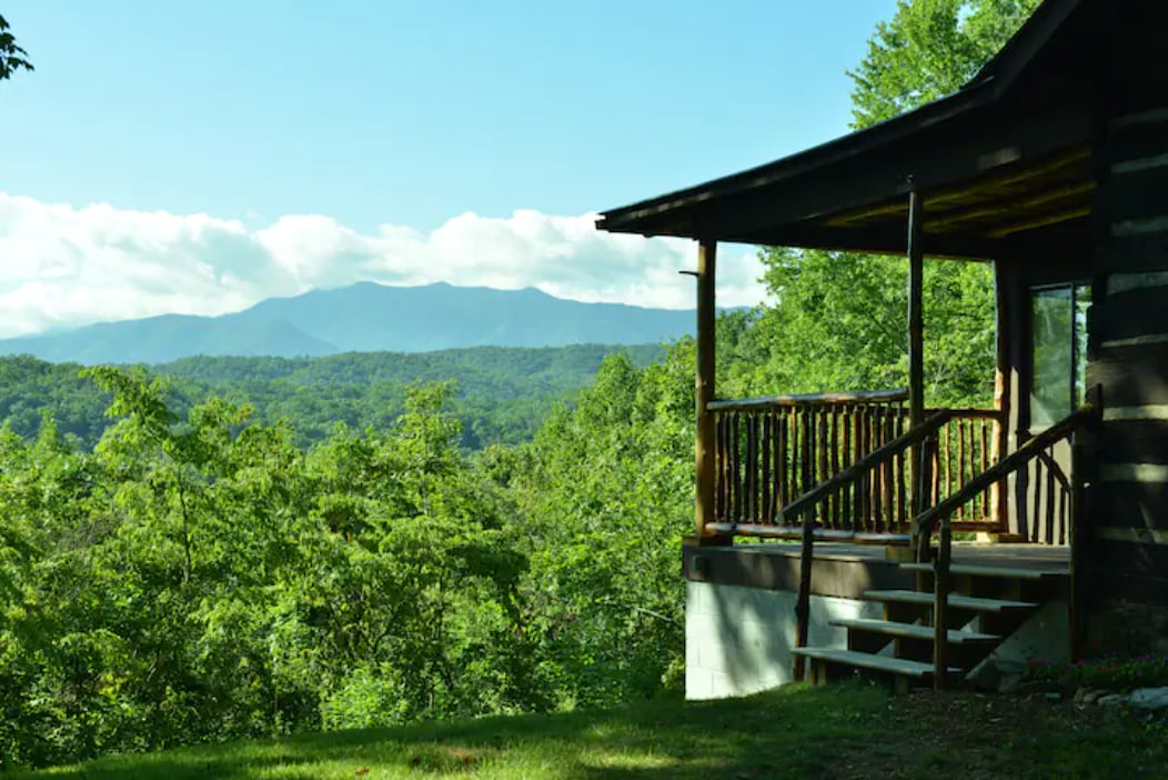 The cabin's porch features wooden railings and steps leading down to a lush yard. A view of the Smoky Mountains is visible in the background, framed by trees that surround the property, providing a sense of tranquility.