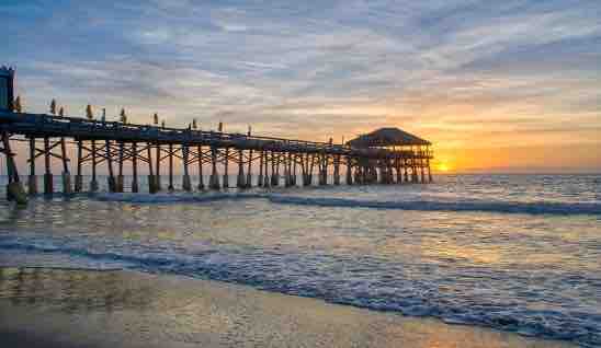 A wooden pier extends into the ocean, silhouetted against a vibrant sunset. Gentle waves lap against the shore, reflecting the warm colors of the sky. The structure is lined with individuals enjoying the view as the sun descends on the horizon.