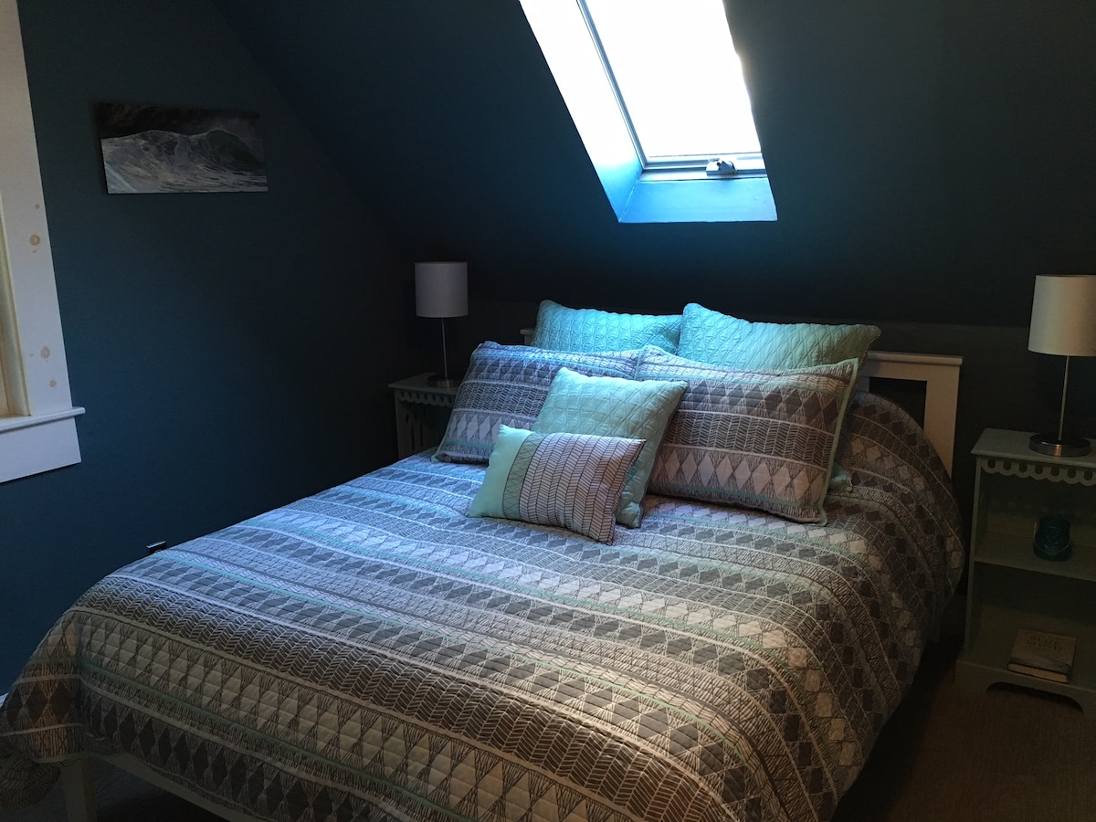 A cozy sleeping area features a spacious queen bed adorned with a textured patterned blanket and multiple accent pillows. Soft blue walls complement the natural light streaming through a skylight above. Nightstands with lamps add functional decor to the inviting space.
