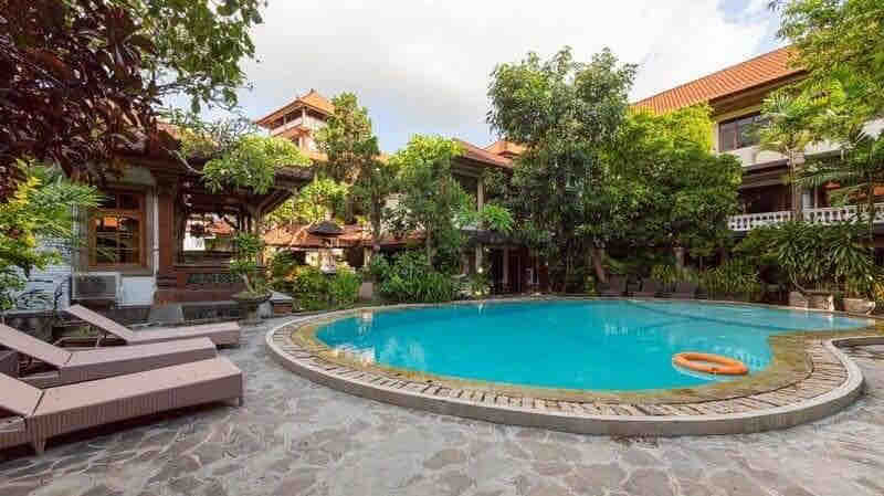 An outdoor swimming pool is surrounded by lush greenery and tropical plants, providing a serene atmosphere. Sun loungers are positioned along the pool's edge, inviting relaxation. The pool's clear blue water contrasts with the stone and tiled decking.