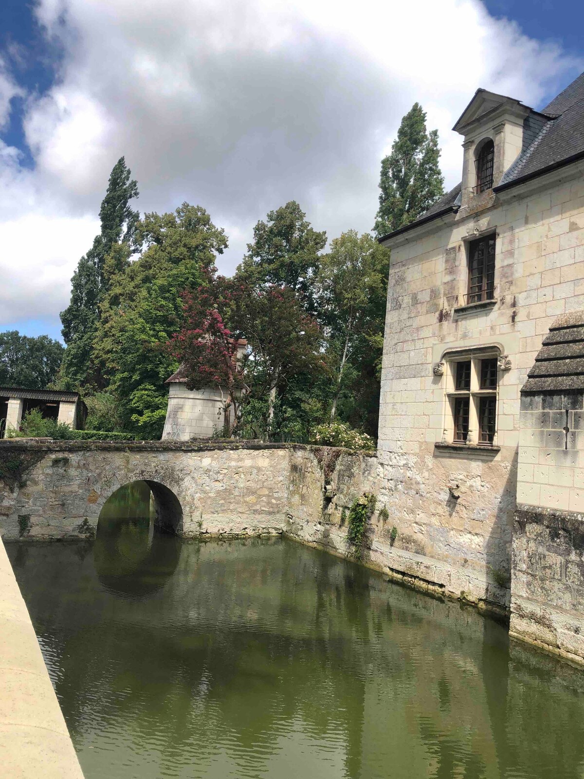 The historic manor is partially surrounded by tranquil water, with a stone bridge leading to the entrance. Towering trees provide a lush backdrop, and the architecture is highlighted by intricately designed windows and a textured stone facade.