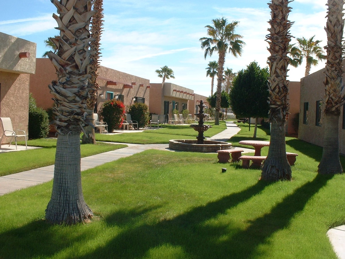 A landscaped courtyard is featured, with paths winding through vibrant green grass. Palm trees provide shade alongside a fountain and seating areas, creating a tranquil environment within the resort. Warm sunshine brightens the adobe-style buildings in the background, enhancing the inviting outdoor space.